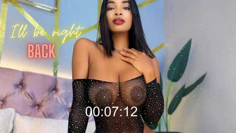 Media: Video of a dark-skinned woman with long, straight black hair, wearing a sheer, sparkly black top, standing in a modern, brightly lit room with a large pink tufted headboard. Text reads, \"I'll be right back\" and \"00:07:12\" in the image.