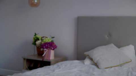 Media: Video of a minimalist bedroom featuring a light gray upholstered headboard, a white bed with two white pillows, a wooden nightstand with a potted plant, and a pendant light hanging above.