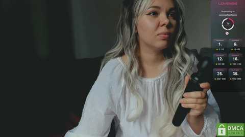 Media: Video of a woman with long, wavy platinum blonde hair, wearing a white blouse, holding a smartphone, looking concerned. Background shows a dimly lit room with a TV displaying weather information.