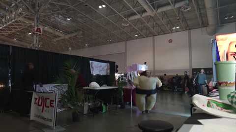 Media: A video of an indoor convention hall with exposed ceilings and industrial lighting. People, including a woman in a green costume, walk around various booths, tables, and banners.