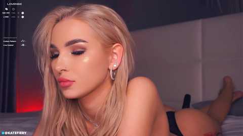 Media: A video of a blonde woman with fair skin, wearing dramatic makeup, lying on a bed with a grey headboard, looking downward. She wears large hoop earrings and is topless.