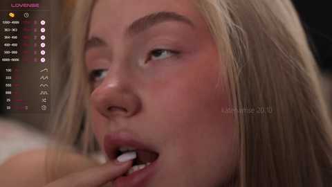 Media: A close-up video of a young, light-skinned woman with long blonde hair, wearing makeup, biting her lip while looking to the left. The background includes a streaming platform with metrics and a watermark.