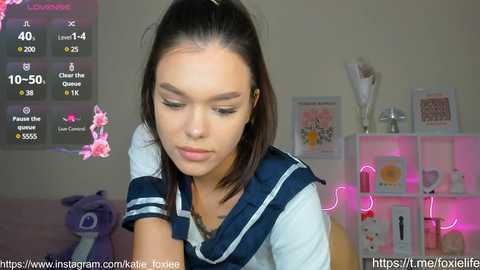 Media: Video of a young woman with light skin, dark hair, and a Japanese schoolgirl outfit, looking down. Background features pink neon lights, framed artwork, and a stuffed toy.