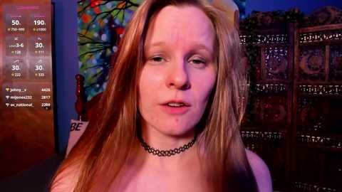 Media: Video of a young woman with long red hair, wearing a black choker necklace, indoors with a colorful stained glass window and a wooden screen in the background.