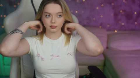 Media: Video of a fair-skinned woman with shoulder-length blonde hair, wearing a white top with floral embroidery, adjusting her hair in a modern room with purple and green lighting and a white chair.