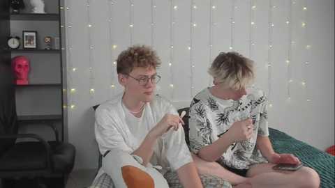 Media: Video of two young men, one with curly hair, glasses, and a white robe, the other in a floral shirt, sitting on a bed, chatting.