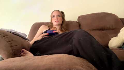 Media: A video of a young woman with light skin and long, light brown hair in a ponytail, wearing a black shirt and pants, sitting on a brown couch playing a video game, with a stuffed bear beside her.