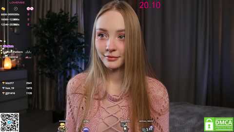 Media: Video of a young Caucasian woman with long, straight blonde hair, wearing a pink knitted sweater, smiling slightly. Background features a dark room with a potted plant and a bed.