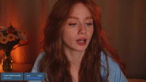 Media: Video of a young woman with long, wavy red hair and pale skin, wearing a light blue top, speaking into a microphone against a warm, softly lit background.