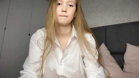 Media: A video of a young Caucasian woman with long, straight blonde hair and fair skin, wearing a white button-down shirt, sitting on a bed with pink pillows and a grey headboard in a modern, minimalist bedroom.