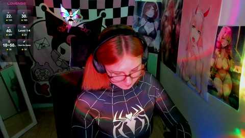 Media: Video of a young woman with red hair, wearing a Spider-Man t-shirt, glasses, and headphones, seated in a dimly lit room with anime posters and a black-and-white checkered wall.