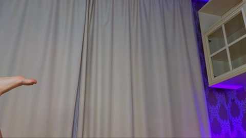 Media: Video of a person's hand holding a gray curtain, revealing a patterned wall and a glass-paneled cabinet with a purple light, creating a dramatic indoor setting.