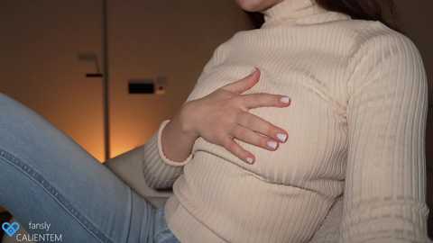 Media: Video of a woman with light skin and straight, brown hair, wearing a beige ribbed turtleneck sweater and blue jeans, touching her breast. Background shows a dimly lit room with a thermostat.