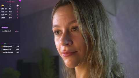 Media: Video of a young woman with long, wavy, light brown hair, fair skin, and brown eyes. She has a soft, neutral expression. The background shows a dimly lit room with a blurry potted plant.