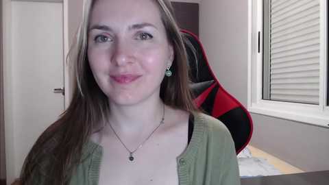 Media: A video of a young woman with light skin, long brown hair, and green eyes, wearing a green cardigan and necklace, smiling, sitting in a room with a black and red gaming chair, white window blinds, and a white door.