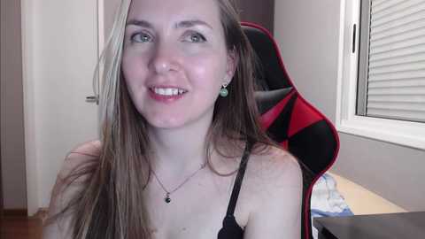 Media: Video of a young Caucasian woman with fair skin and long, straight blonde hair, wearing a black bra and a necklace, sitting on a red and black gaming chair in a brightly lit room.