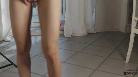 Media: Video of a nude woman standing in a bright, tiled room with white curtains. She has fair skin, slender legs, and a small waist.