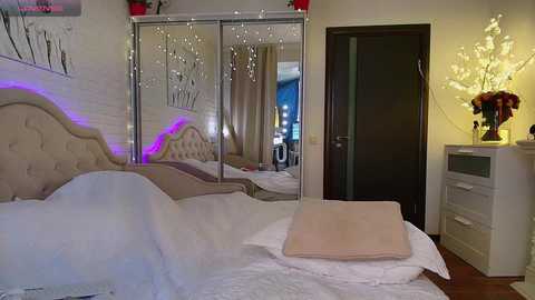 Media: A modern bedroom with a tufted beige headboard, white bed, and mirror reflecting a lit vanity. A dark wooden door, white dresser, and floral arrangement adorn the space.