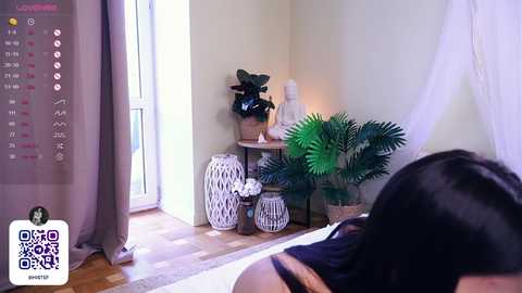 Media: A video of a serene bedroom corner featuring a woman with long black hair, a white nightstand with a plant, and a calendar.