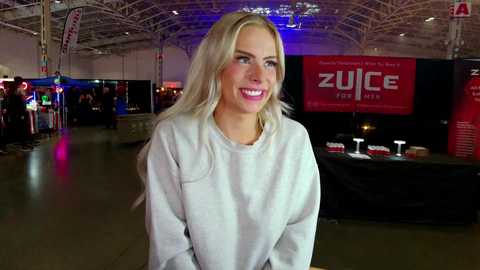 Media: Video of a smiling, fair-skinned, blonde woman in a white sweater, standing in a brightly lit, modern warehouse with \"ZUCCHERI\" signage, and colorful LED lights.