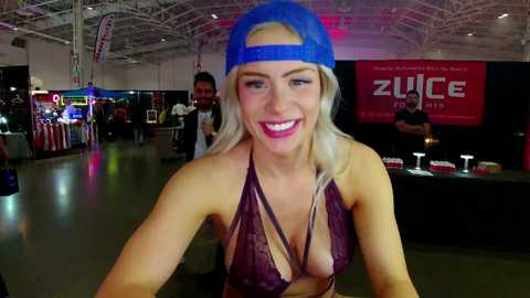 Media: Video of a smiling, blonde woman in a blue backwards baseball cap, maroon lace-up bikini top, and high heels, posing in a dimly lit convention hall with neon lights and vendor booths in the background.