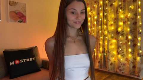 Media: Video of a young woman with long, straight brown hair, wearing a white strapless top, standing in a warmly lit room with yellow string lights and floral curtains.