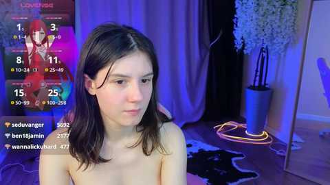 Media: Video of a fair-skinned, slender young woman with shoulder-length dark hair, wearing a red bra, standing in a dimly lit room with a cowhide rug, neon lights, and a potted plant.