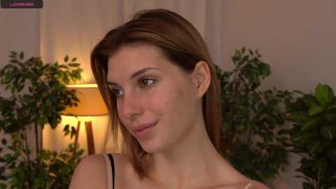 Media: Video of a Caucasian woman with light brown hair, wearing a gray top, smiling slightly, in a room with a lit lamp, green plants, and neutral-colored walls.