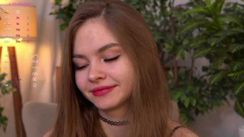 Media: Video of a young woman with long brown hair, closed eyes, and a serene expression, wearing a choker necklace. Background features a green plant and a lamp with a colorful shade.