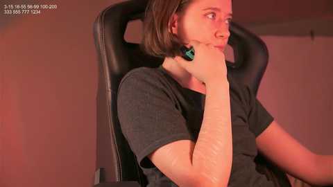 Media: Video of a woman with shoulder-length brown hair, wearing a black t-shirt, seated in a black gaming chair, appearing contemplative with hand to chin, in a dimly lit room.