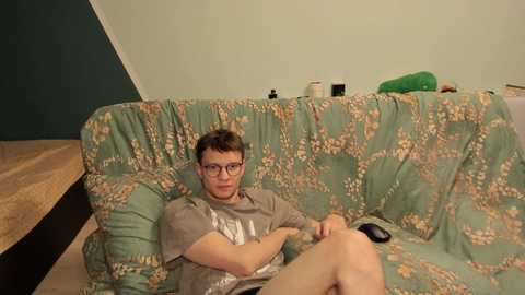 Media: Video of a young man with glasses and short brown hair, wearing a gray t-shirt and shorts, sitting on a floral-patterned couch in a dimly lit room with green walls and a wooden table.