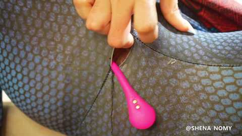 Media: A close-up video shows a person's hand pulling a bright pink vibrator out of a tight, textured gray-blue bodysuit. The image has a watermark \"\u00a9SheNaNomY\" in the bottom right corner.