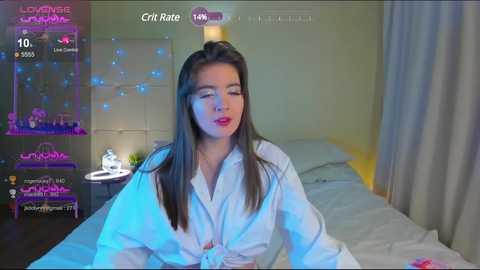 Media: Video of a young woman with long brown hair, wearing a white robe, sitting on a bed in a dimly lit bedroom, with digital overlays of \"LoveRival\" and \"Crist Rate\" in the top corners.