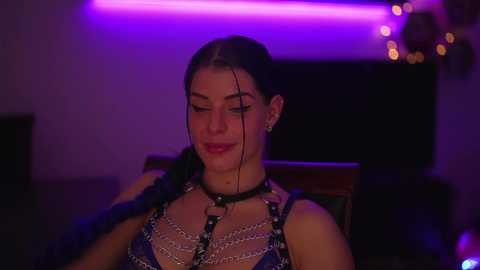 Media: Video of a woman with dark hair and fair skin, wearing a black leather harness, smiling, set against a dimly lit room with purple neon lights.