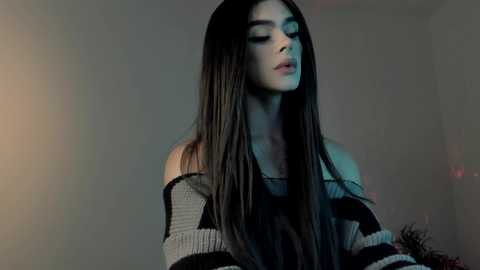 Media: Video of a young woman with long, straight, dark hair, wearing a loose, off-shoulder, grey and black striped sweater, looking contemplative against a dim, neutral background.