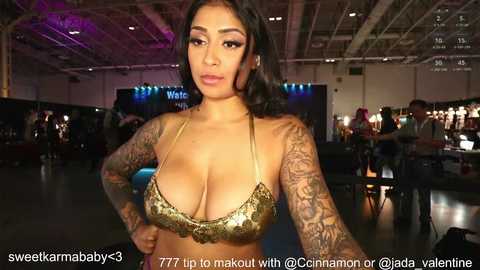 Media: Video of a curvy, tattooed, light-skinned woman with large breasts wearing a gold sequin bikini top, taken in a dimly lit, industrial warehouse.