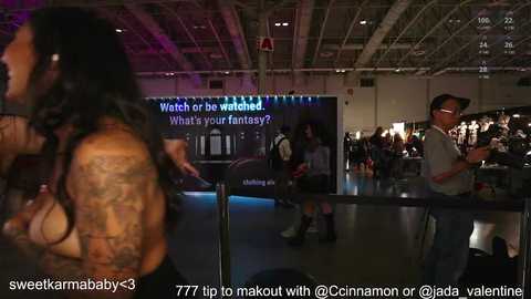 Media: Video of a tattooed woman in a black top and long hair standing in a dimly lit bar with a screen displaying \"What's your fantasy?\" and a man in glasses behind a bar counter.