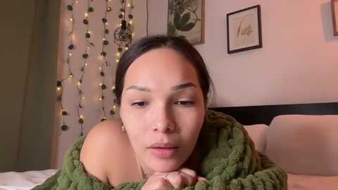 Media: Video of a young woman with medium brown skin and dark hair, wearing a green knit sweater, lying on a bed with white linens. The background features string lights and framed botanical prints on a light-colored wall.