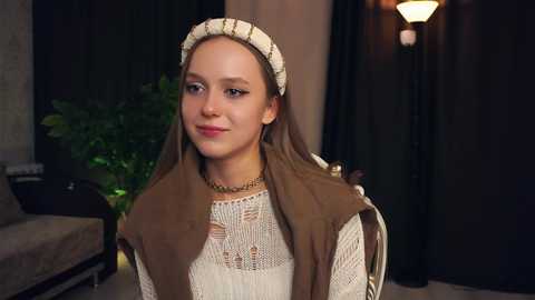 Media: Video of a young girl in a medieval-style costume with a beige headband, brown dress, and intricate white lace. She stands indoors, with dark curtains, a potted plant, and a lamp in the background.