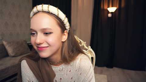 Media: Video of a young woman with fair skin and straight, shoulder-length brown hair, wearing a beige lace dress, a gold headpiece, and a gold necklace, smiling softly, in a dimly-lit room with beige wallpaper and dark curtains.