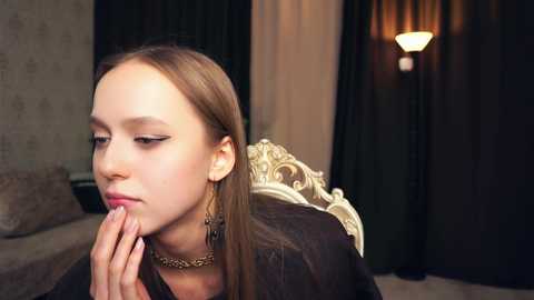 Media: Video of a young Caucasian girl with straight, light brown hair, wearing black clothing and a gold necklace, praying with hands clasped, in a dimly lit room with dark curtains and a soft lamp.