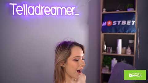 Media: Video of a young, fair-skinned woman with blonde hair, smiling, in a modern, dimly lit room with a purple neon sign reading \"Telldreamer,\" a blue bookshelf with a \"Mostbet\" pillow, and a green \"LuckyBaba\" logo.