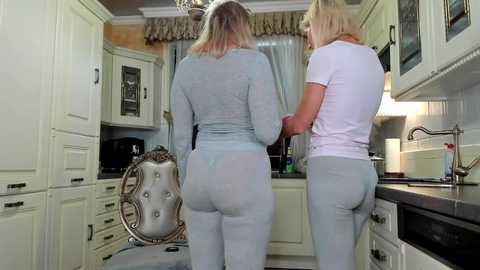 Media: Video of two women in light blue pajamas, standing in a cozy, vintage-style kitchen with cream cabinets, a chandelier, and a tufted chair.