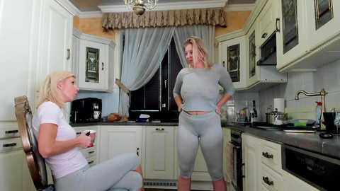 Media: Video of two blonde women in matching light grey athletic outfits, one seated, one standing in a modern kitchen with white cabinets, stainless steel appliances, and a window with sheer curtains.