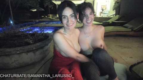 Media: Video of two topless women, one in a red dress, sitting closely on a patio by a pool at night, smiling.