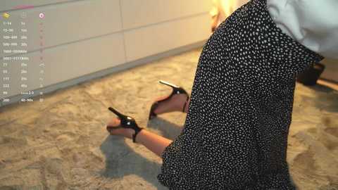 Media: Video of a person in a black polka dot skirt, kneeling on a beige carpet, holding black high-heeled shoes. Background features a white dresser and a lit candle.