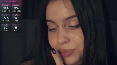 Media: Video of a young woman with long black hair and light skin, smiling slightly, wearing a nose ring. Background includes a black curtain and a digital screen displaying live stream information.