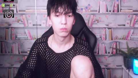 Media: Video of a young Asian man with short black hair, wearing a black mesh shirt, sitting in a chair with a potted plant in the background, surrounded by bookshelves.