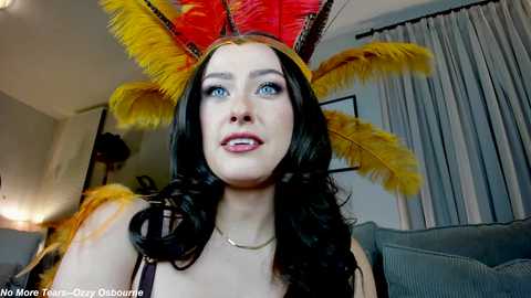 Media: Video of a fair-skinned woman with long, dark hair, wearing a bright red and yellow feathered headdress and a black top, in a modern, dimly lit living room with grey curtains.