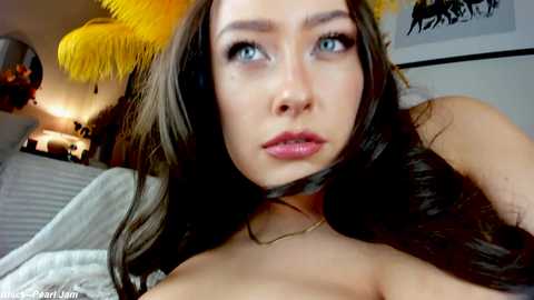 Media: Video of a young woman with long brown hair, blue eyes, and fair skin, wearing a choker necklace. She has a neutral expression, looking slightly to the side. Background includes a yellow feather lamp and framed art.